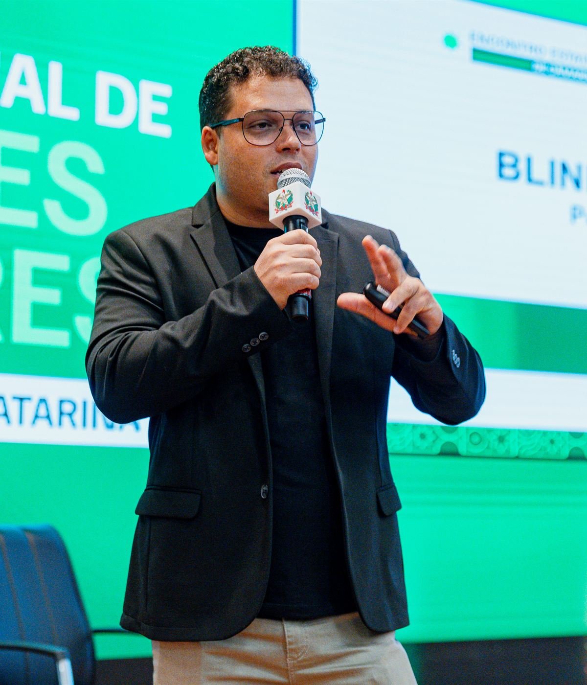 Elias Edenis no palco, de blazer, falando ao microfone diante de um telão com o seu nome.