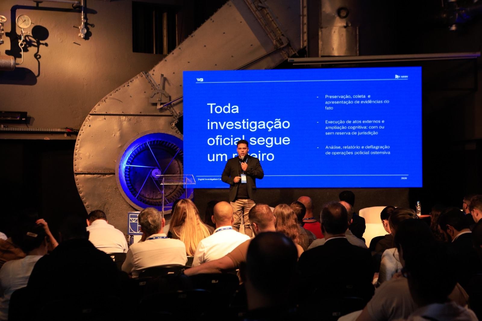 Auditório cheio visto do fundo; Elias Edenis no palco diante de um telão azul com o texto “Toda investigação oficial segue um roteiro”.